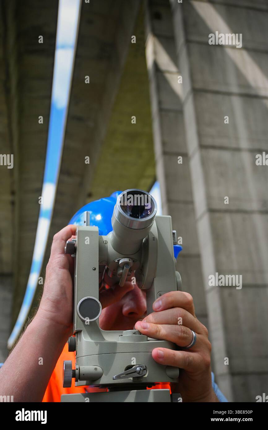 Surveyor che lavora con apparecchiature di transito teodolite in cantiere stradale sotto un ponte di cemento. Foto Stock