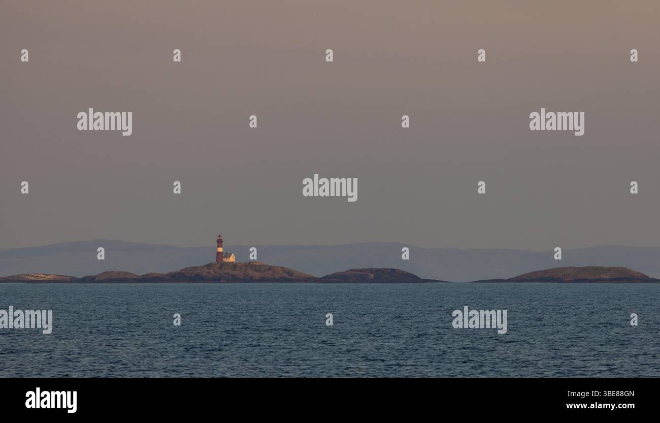 Faro remoto dell'isola al largo della costa della Norvegia, illuminato dalla luce soffusa della sera sotto un cielo pastello, visto attraverso il mare calmo. Foto Stock