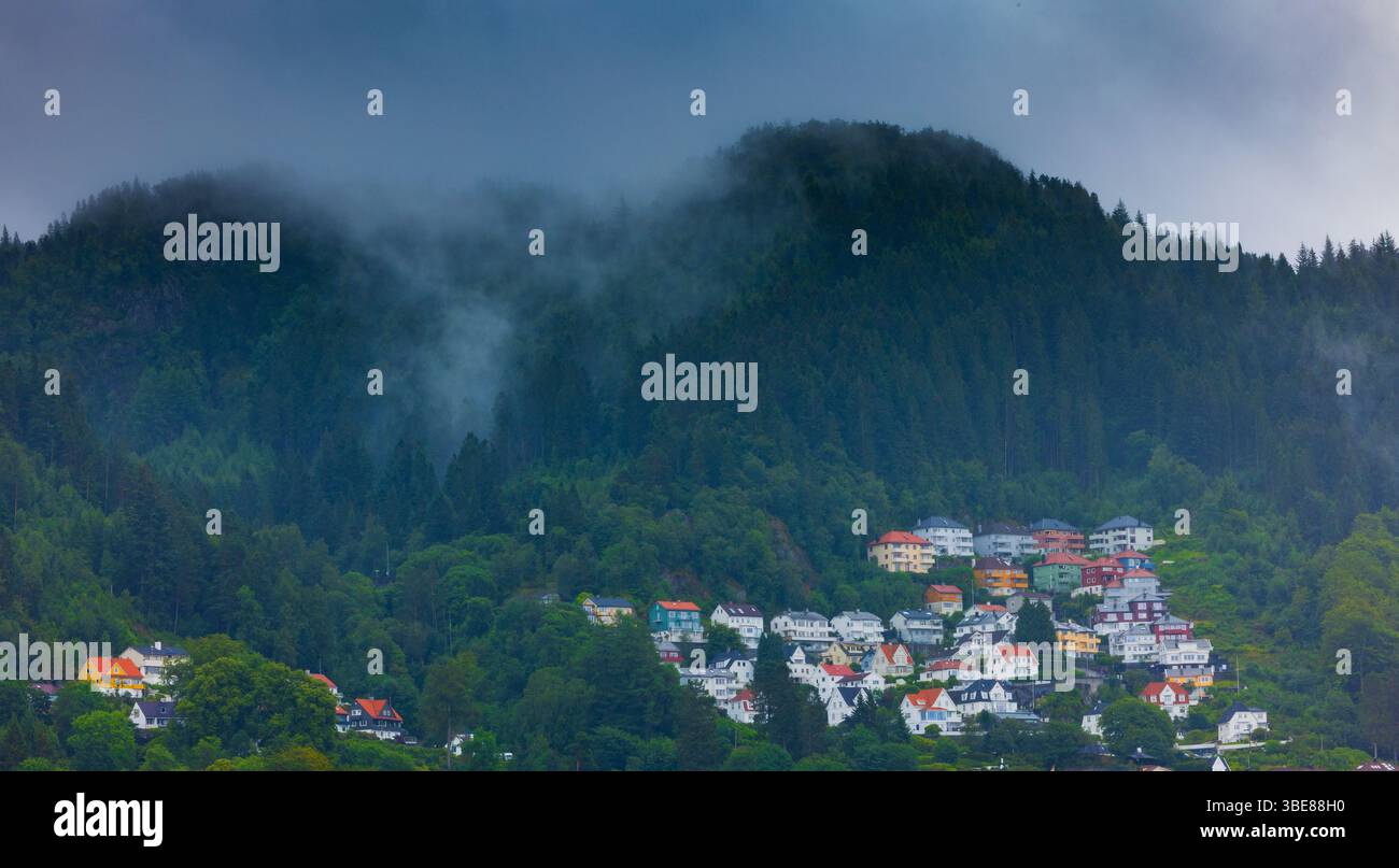 Case colorate accoccolate ai piedi delle colline boscose coperte di nebbia in Norvegia, catturate durante il tempo coperto. Foto Stock