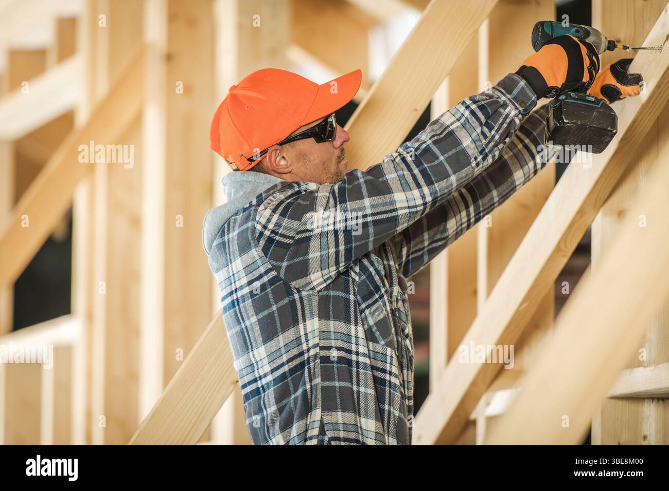 Un operaio edile che indossa attrezzature protettive perfora le travi di legno in un cantiere, mostrando abilità artigianale e concentrandosi sulla sicurezza. Foto Stock