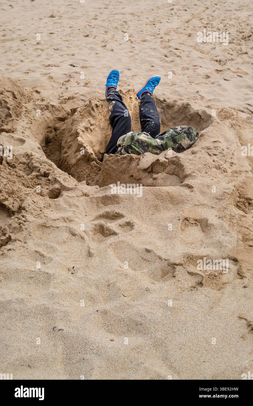 Un giovane seduto e addormentato in un buco scavato nella sabbia su una spiaggia di Newquay, in Cornovaglia, nel Regno Unito. Foto Stock
