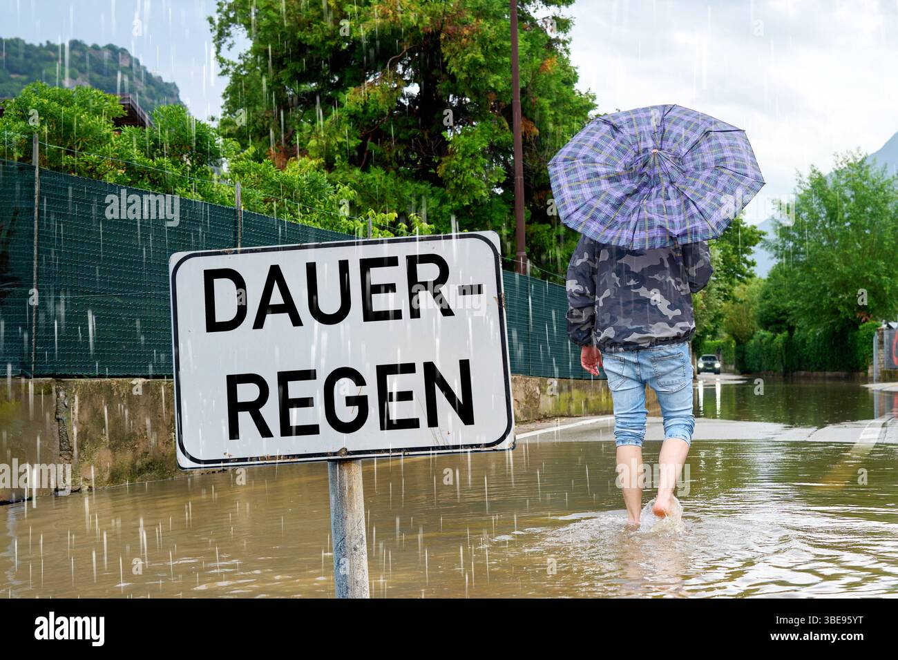 Bavaria, Germania - 28 maggio 2025: FOTOMONTAGGIO, Un uomo con un ombrello cammina attraverso una strada allagata - il cartello Dauerregen simboleggia la situazione meteorologica in Baviera con possibili inondazioni locali dovute a persistenti piogge. *** FOTOMONTAGE, Ein Mann mit Regenschirm watet durch eine überflutete Straße das Schild Dauerregen steht sinnbildlich für die Wetterlage in Bayern mit möglichen lokalen Überschwemmungen durch anhaltende Regenfälle. Foto Stock