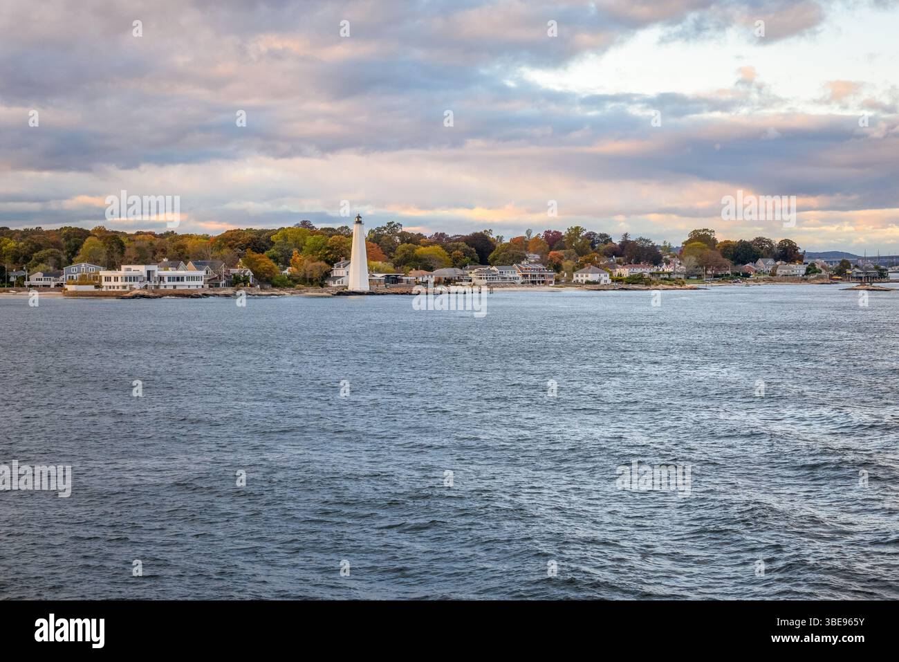 Faro e proprietà sul lungomare su una costa frastagliata delimitata da colorati boschi autunnali a Connceticut sotto il cielo nuvoloso al tramonto Foto Stock