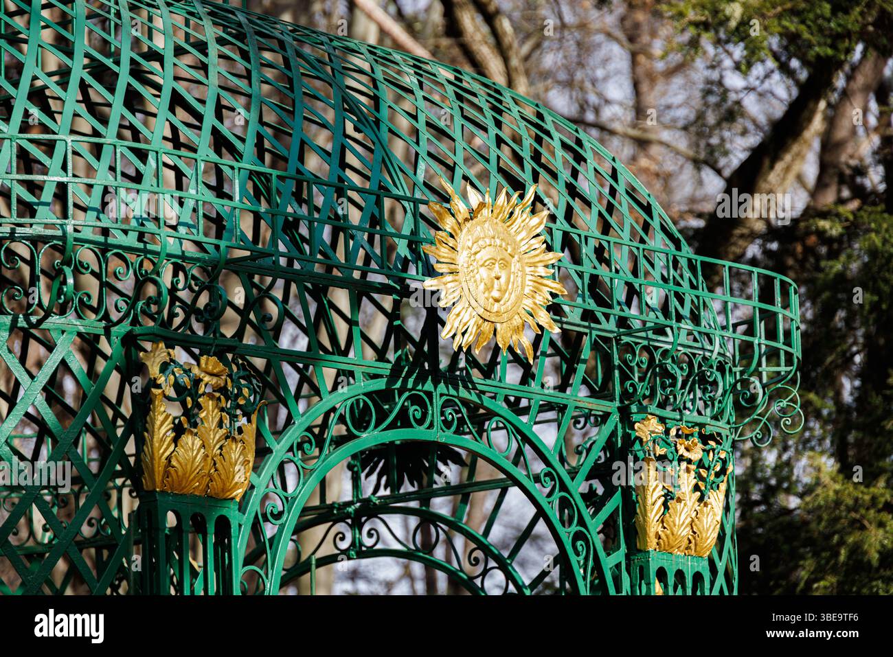 Gazebo decorativo in ferro battuto verde con motivi dorati di sole e fogliame, adagiato su uno sfondo boschivo all'inizio della primavera o nel tardo inverno. Foto Stock