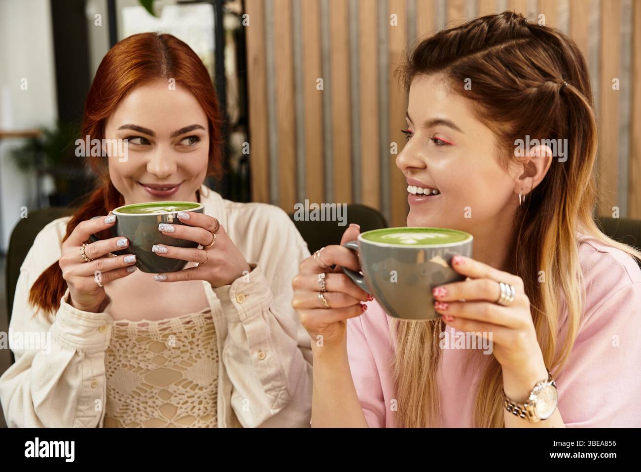 Due giovani donne condividono risate e bevande verdi in un moderno ambiente da caffè, abbracciando l'amicizia. Foto Stock