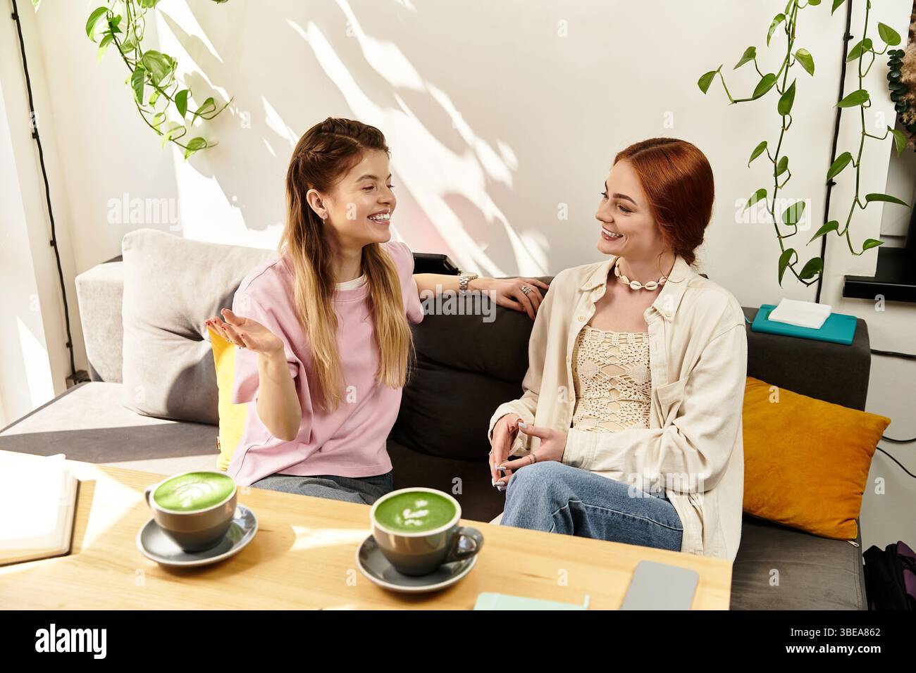 Due giovani donne condividono risate e storie sorseggiando un drink verde in un accogliente caffè. Foto Stock