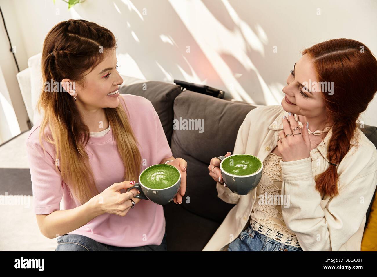 Due giovani donne condividono risate e deliziosi drink verdi mentre si rilassano in un accogliente caffè. Foto Stock