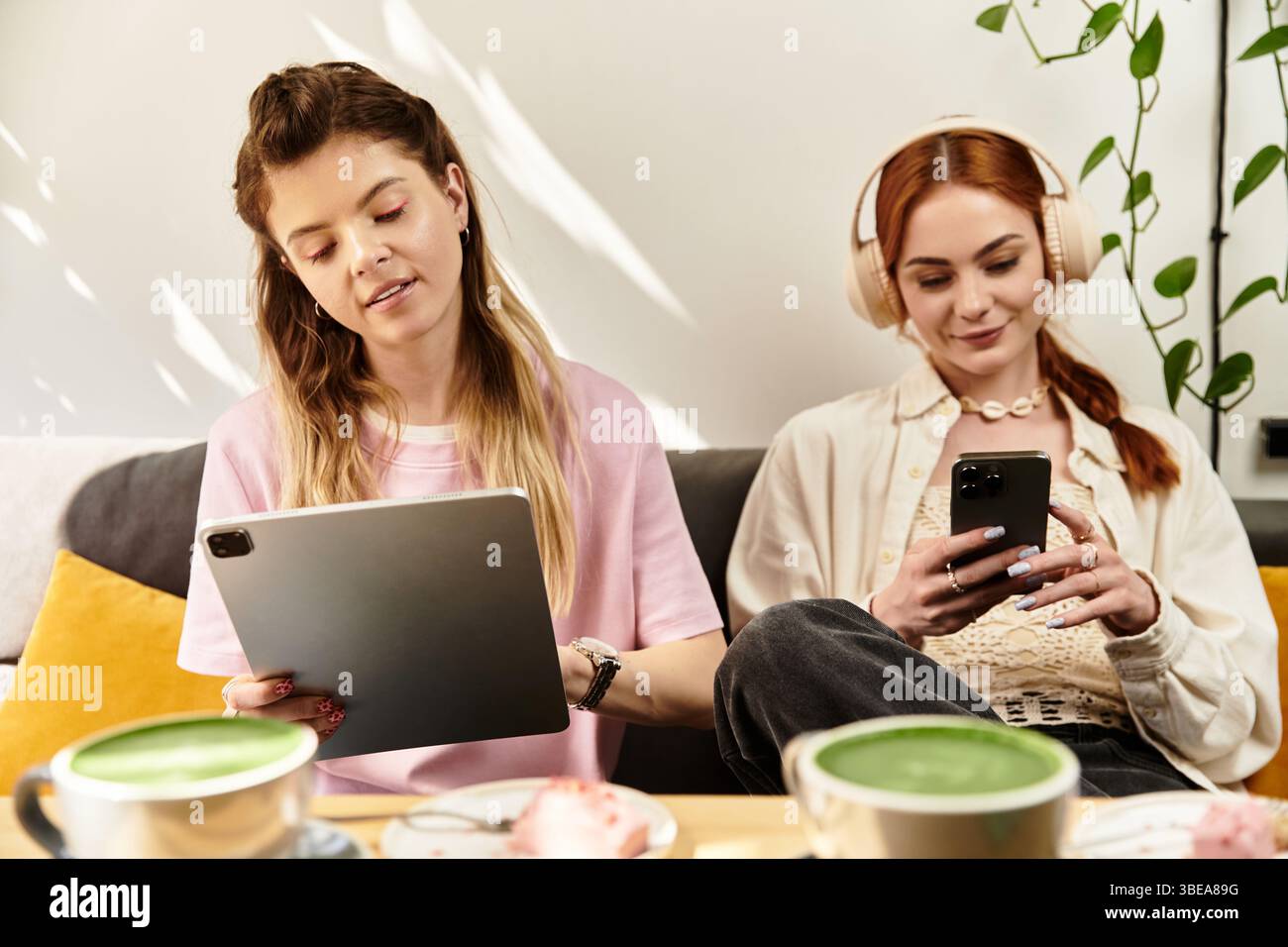 Due amici si rilassano in un elegante caffè, impegnati con i loro dispositivi vicino a bevande verdi. Foto Stock