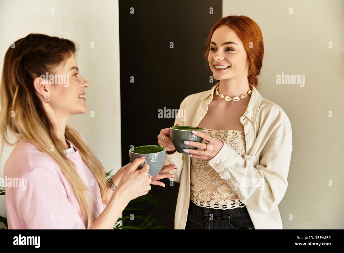 Due giovani donne condividono le risate mentre tengono bevande verdi in un ambiente alla moda. Foto Stock