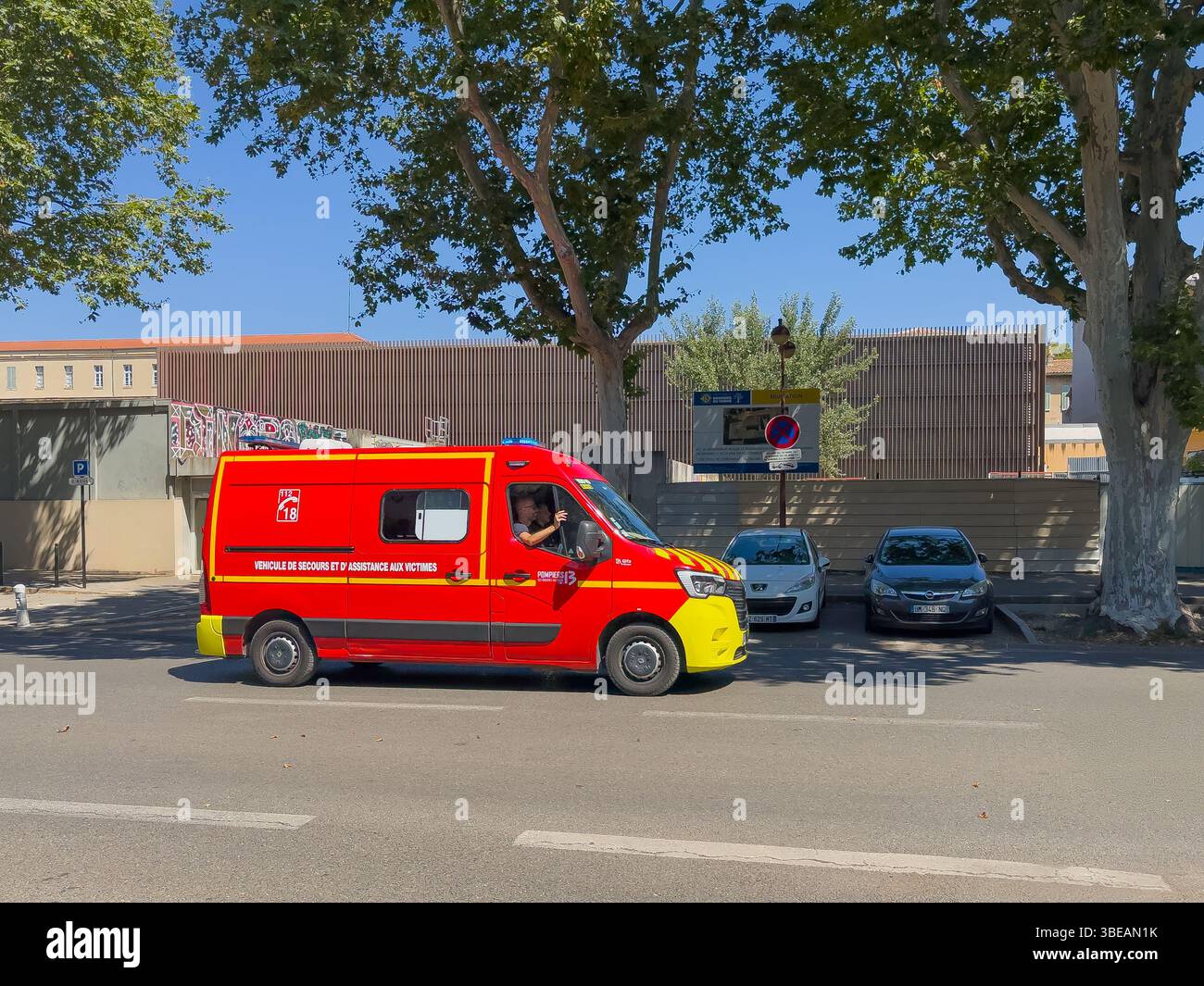 Marsiglia, Francia - 10 luglio 2024: Scenario del centro. Furgone rosso-giallo ambulanza in strada sotto fogliame verde, auto parcheggiate Foto Stock