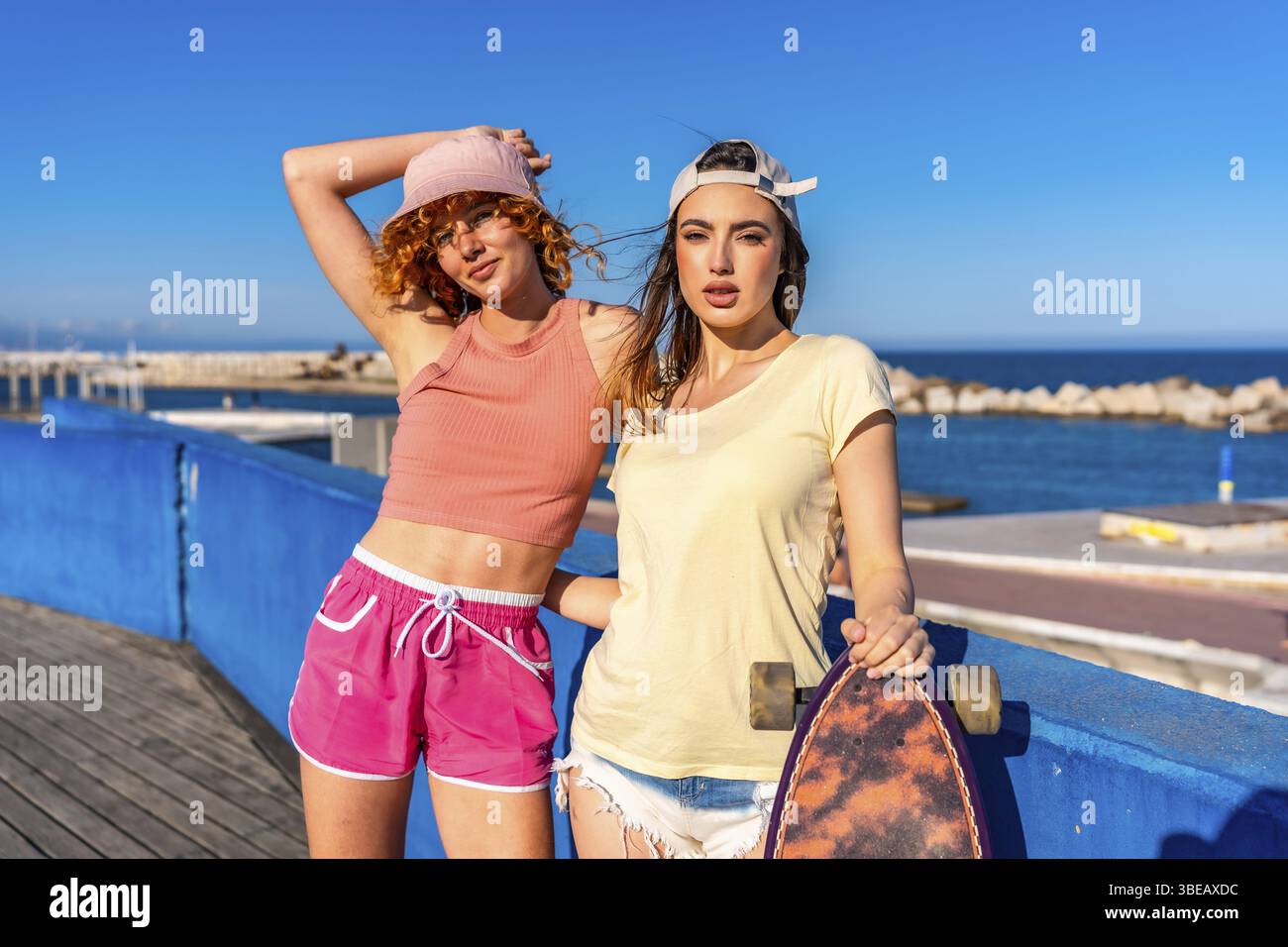 Due giovani donne che si rilassano in riva al mare in una giornata di sole, una con skateboard, incarnano uno stile di vita spensierato e avventuroso Foto Stock