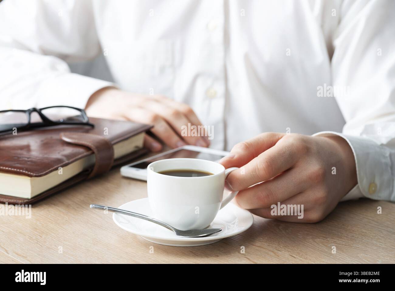 Uomo in camicia bianca che usa lo smartphone seduto alla scrivania con una tazza di caffè. Applicazioni aziendali e tecnologia mobile. Navigazione Internet e comunicazione Foto Stock