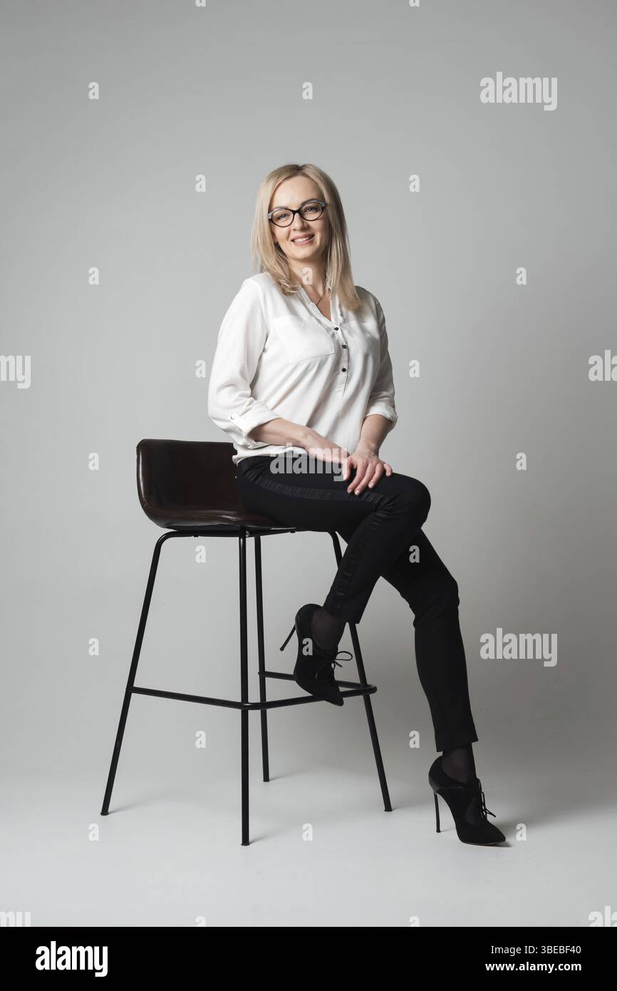 Bella e sorridente giovane donna con occhiali seduti su una sedia in uno studio con sfondo grigio. Agente di vendita, manager, agente, assistente, insegnante, prigioniero Foto Stock