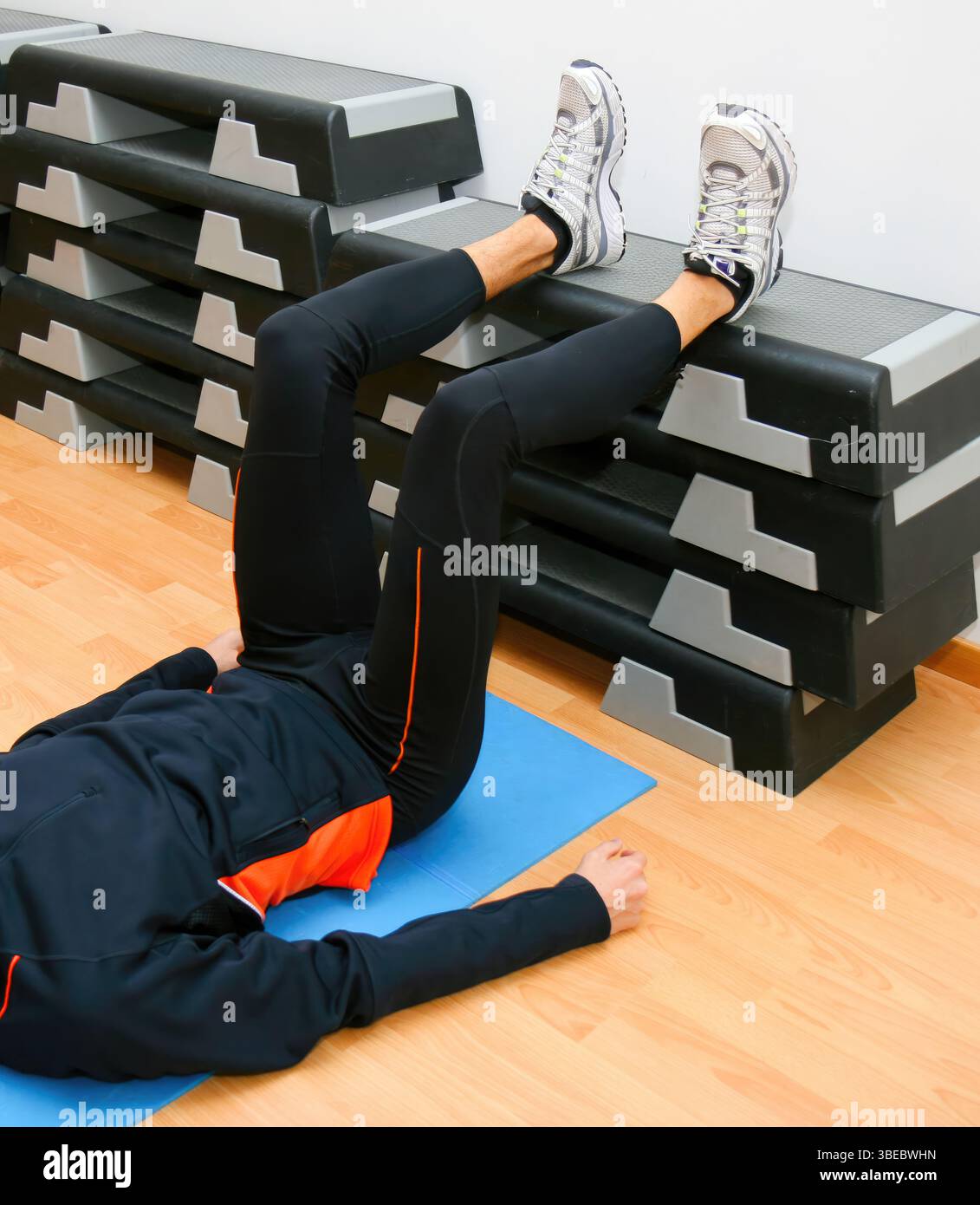 L'atleta giace su un tappetino da palestra, eseguendo un esercizio di sollevamento delle gambe con i piedi appoggiati su uno stepper, dimostrando forza e flessibilità in una routine di fitness Foto Stock