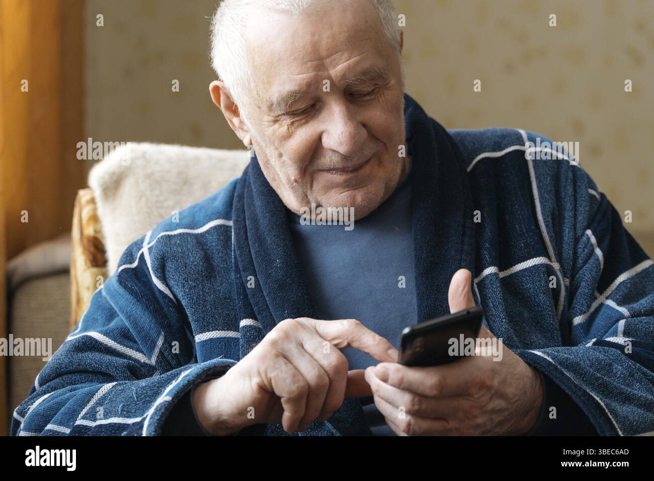 Uomo anziano con smartphone seduto su una poltrona e che scorre sui social media. . Foto Stock