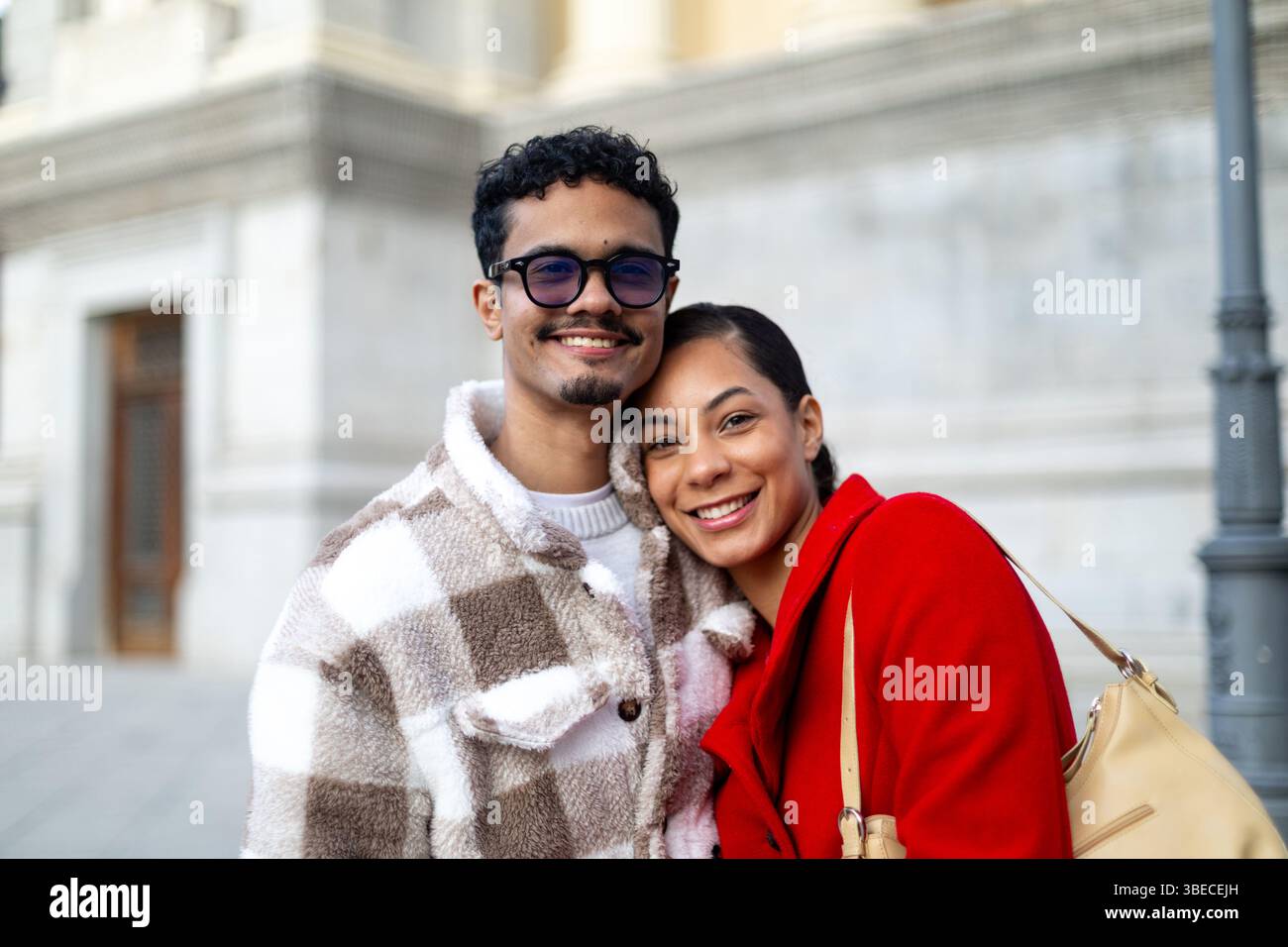 Giovane coppia multietnica che sorride e abbraccia all'aperto in un ambiente urbano, mostrando amore, felicità e solidarietà Foto Stock