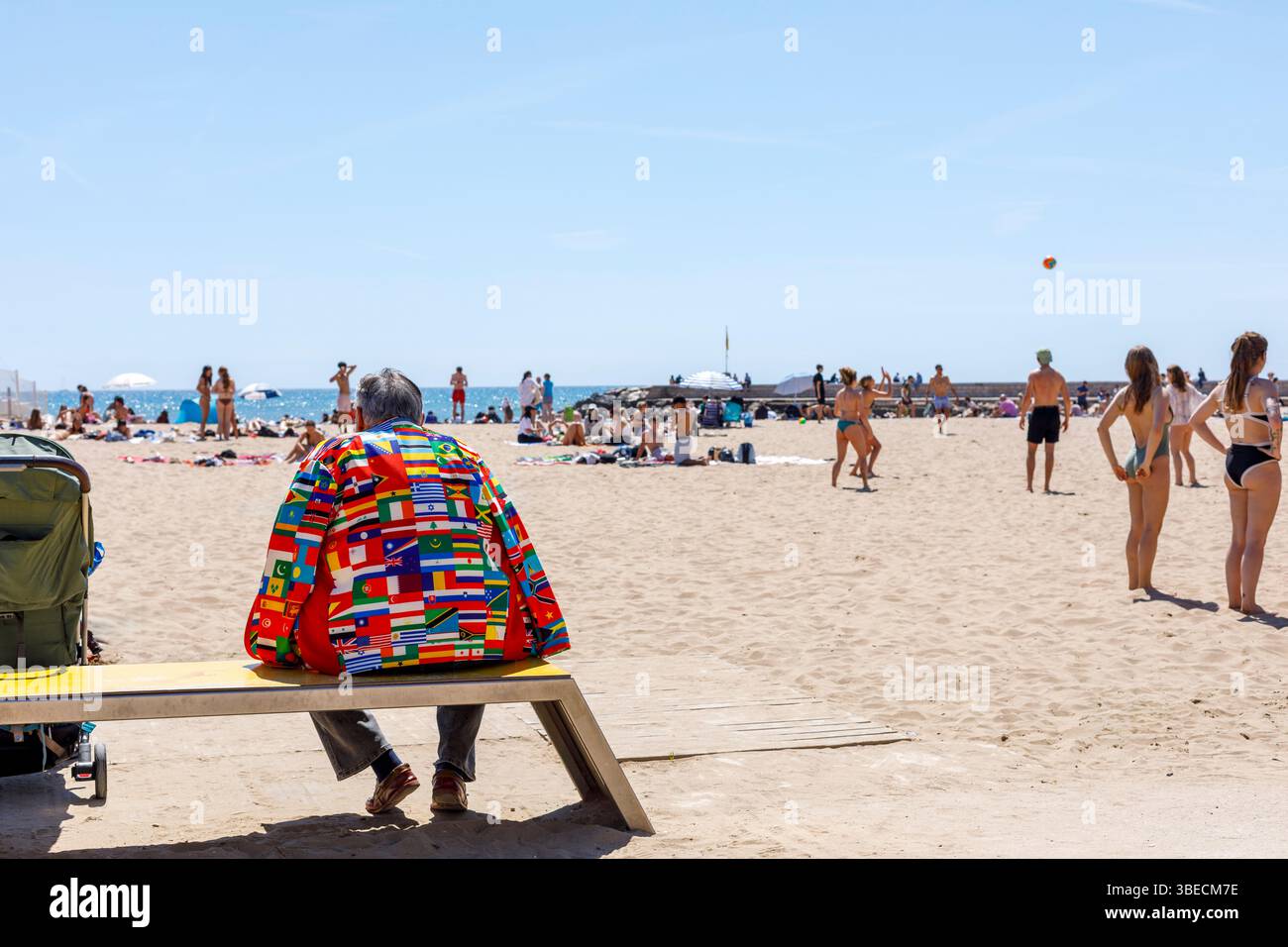 Spagna, Sitges, Un uomo con una giacca fatta di bandiere di tutto il mondo seduto su una spiaggia Foto Stock
