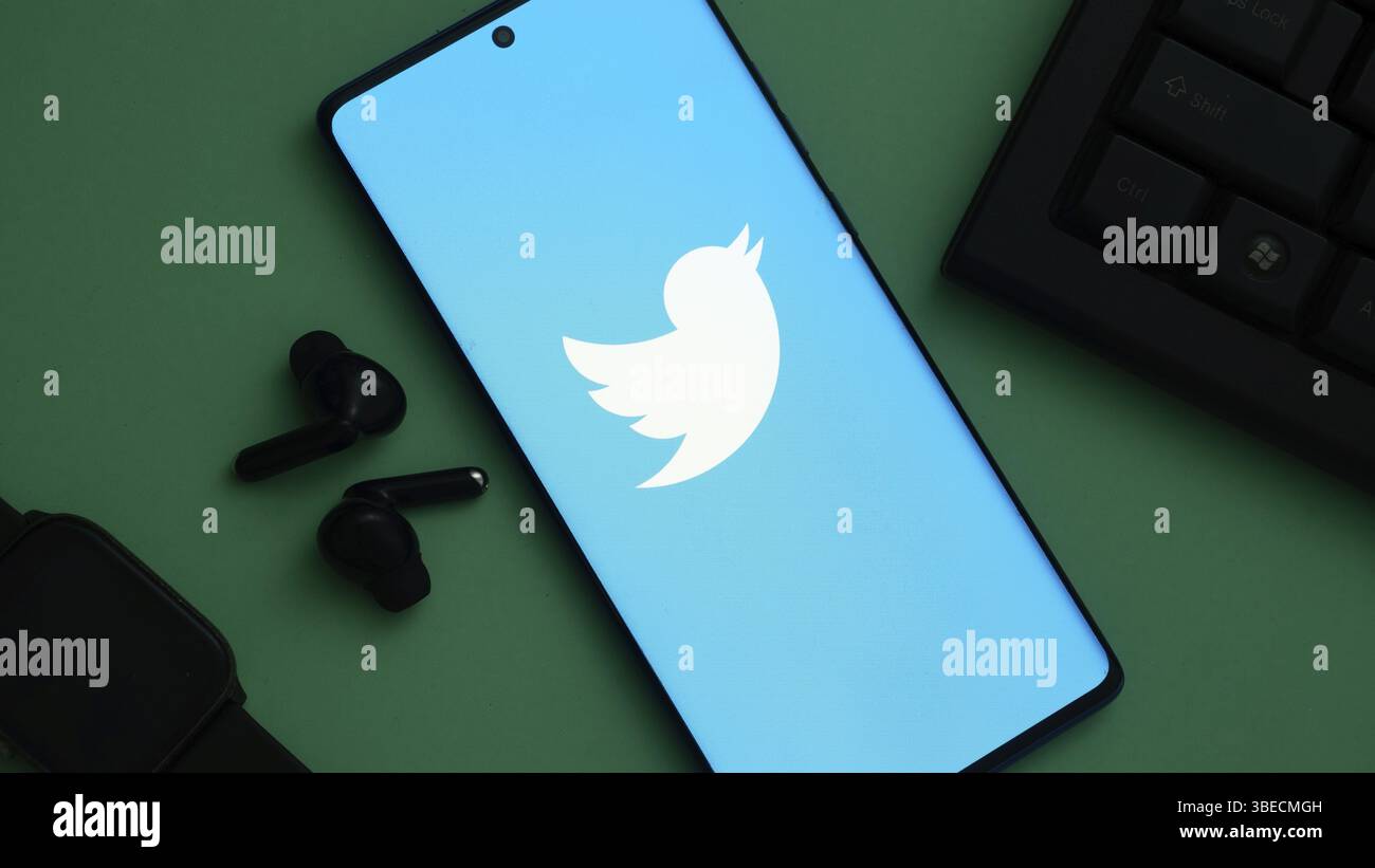 Logo Twitter su smartphone con schermata blu, accanto agli auricolari e alla tastiera neri Foto Stock