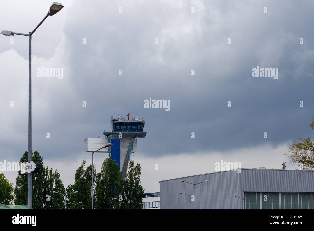 Norimberga, Germania - 25 maggio 2025: Torre di controllo aeroportuale sotto il cielo nuvoloso. Lampioni e fogliame in primo piano. Edificio sulla destra. Foto Stock
