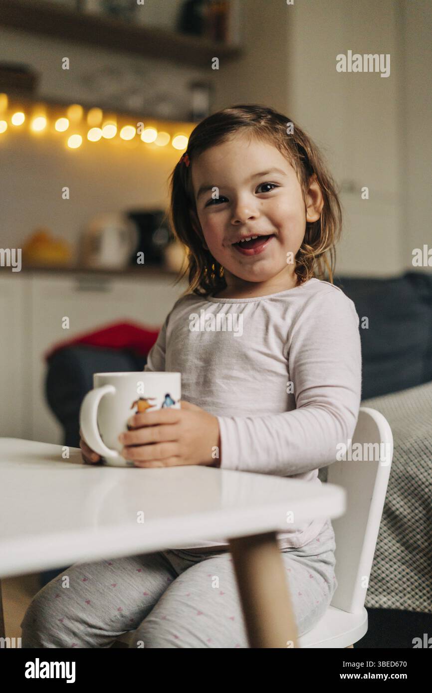 Bambina caucasica che beve cacao dalla tazza a casa. Luci bokeh natalizie. Foto Stock