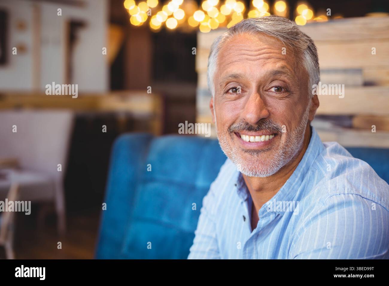 Uomo anziano seduto sulla panchina blu nel bar con una camicia a righe azzurre sotto le luci sospese Foto Stock