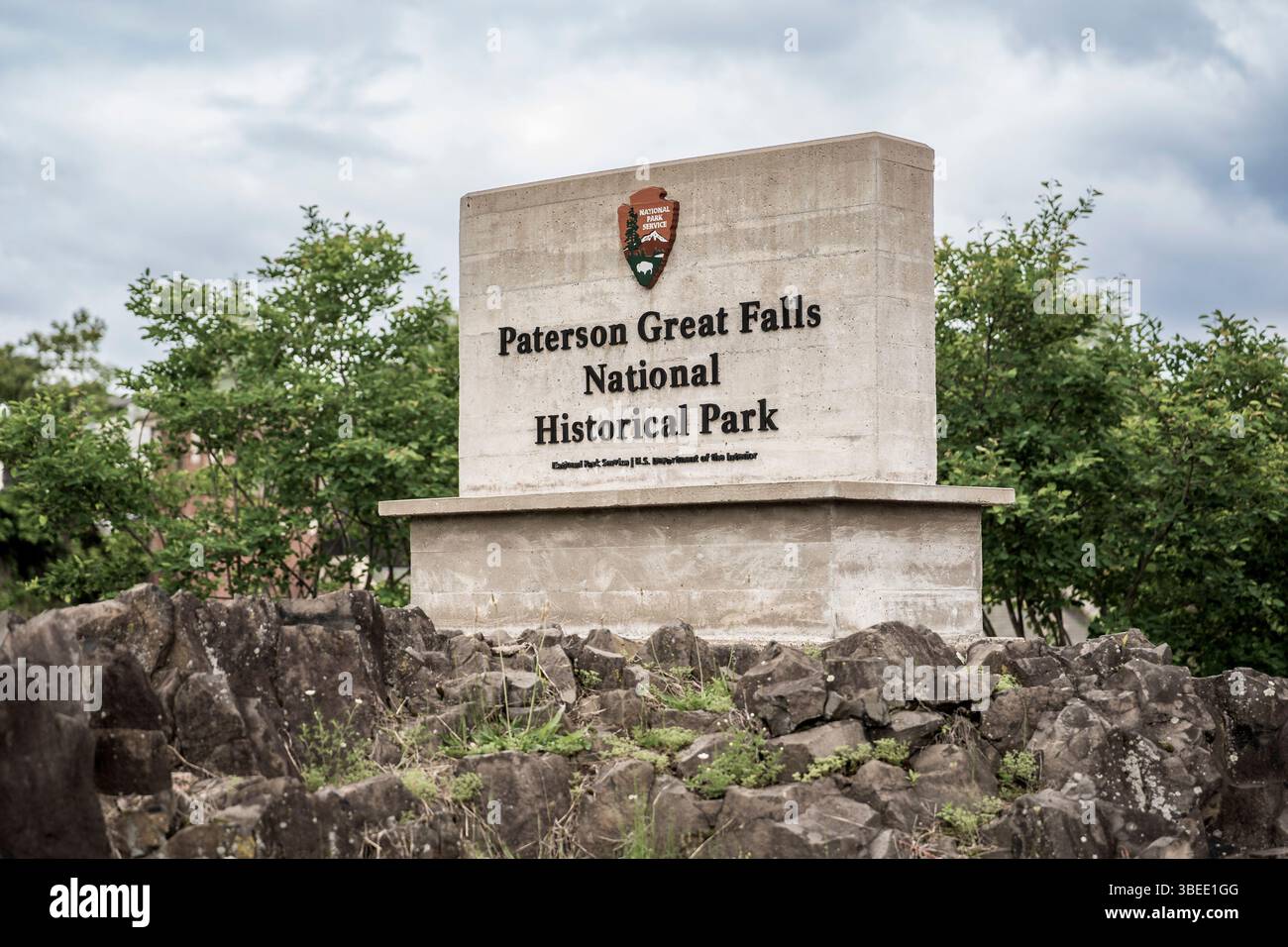 Paterson, New Jersey - Stati Uniti - 24 maggio 2025 cartello di pietra per il Paterson Great Falls National Historical Park si erge tra rocce aspre e vegetazione sotto una nuvolosa sk Foto Stock