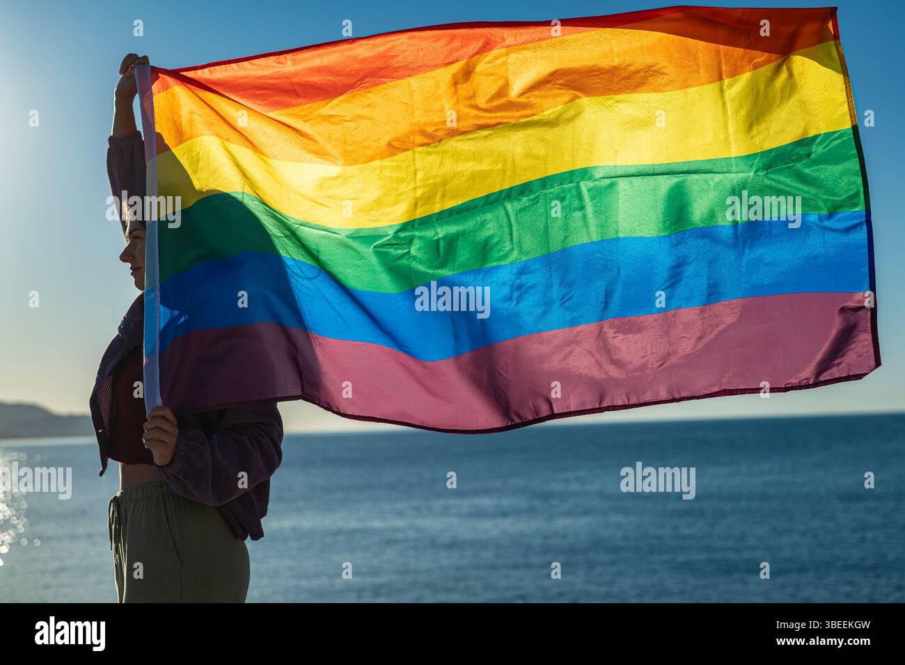 Profilo di una giovane donna che regge una bandiera arcobaleno soffiata dal vento. L'immagine retroilluminata viene scattata dal mare, con la bandiera che occupa la maggior parte del fotogramma. B Foto Stock