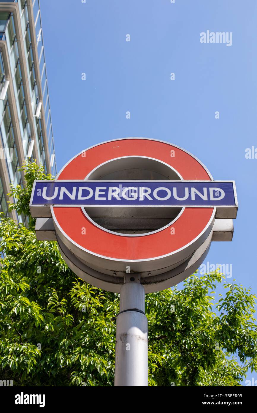Un cartello della metropolitana di Londra si trova in primo piano contro un cielo azzurro. L'iconico design rosso e blu è distinto e riconoscibile nel paesaggio urbano. Foto Stock