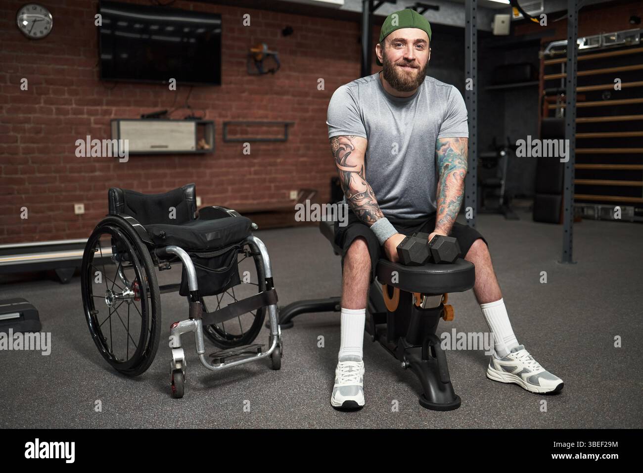 Uomo con tatuaggi che si allena in palestra utilizzando attrezzature adattive, mostrando la sua determinazione e forza, mentre la sedia a rotelle è presente sullo sfondo Foto Stock