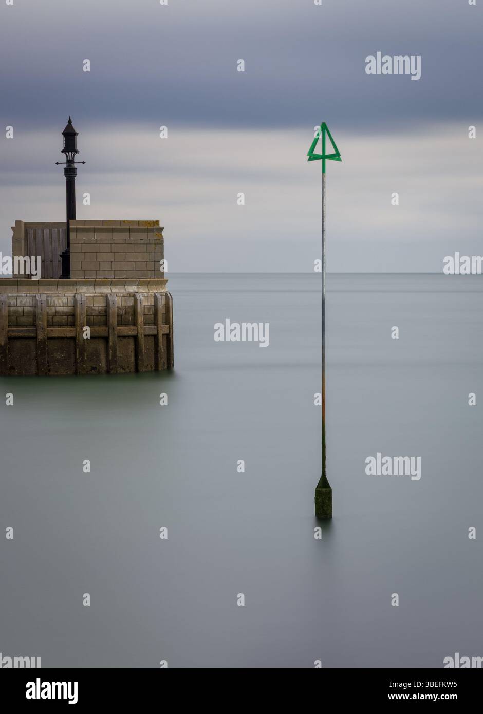 Fotografia a lunga esposizione di un marcatore di canale verde e di una struttura di molo in pietra sotto cieli nuvolosi al largo della costa dell'Isola di Wight, Regno Unito. Foto Stock