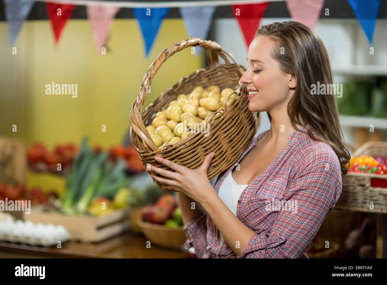 Donna che tiene un cestino di vimini ripieno di patate gialle al mercato con pomodori e bandiere Foto Stock