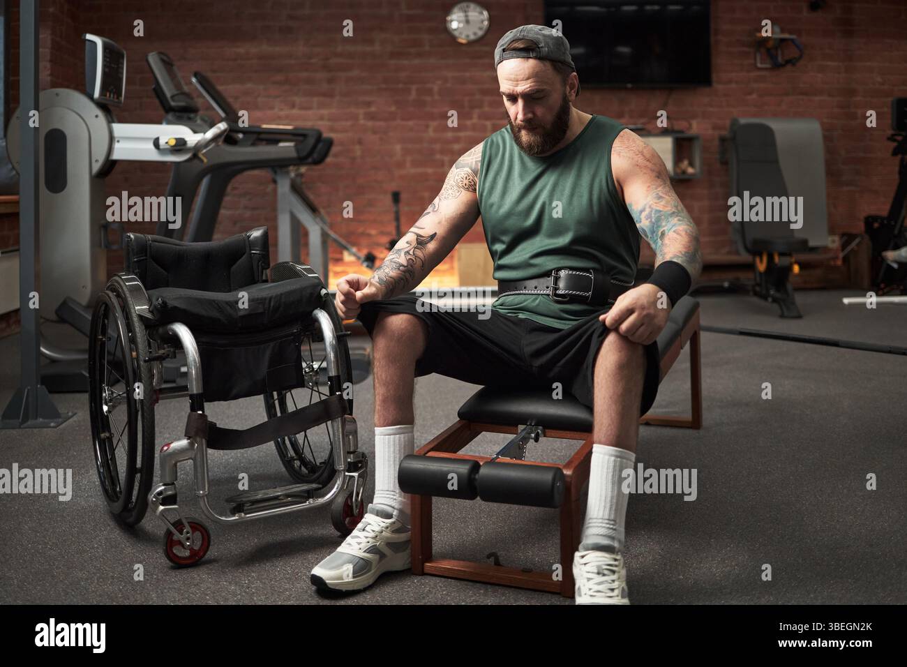 Uomo in palestra che utilizza attrezzature adattive per allenarsi accanto alla sedia a rotelle in background. Si concentra sul percorso di fitness personale senza lasciar perdere i limiti fisici Foto Stock