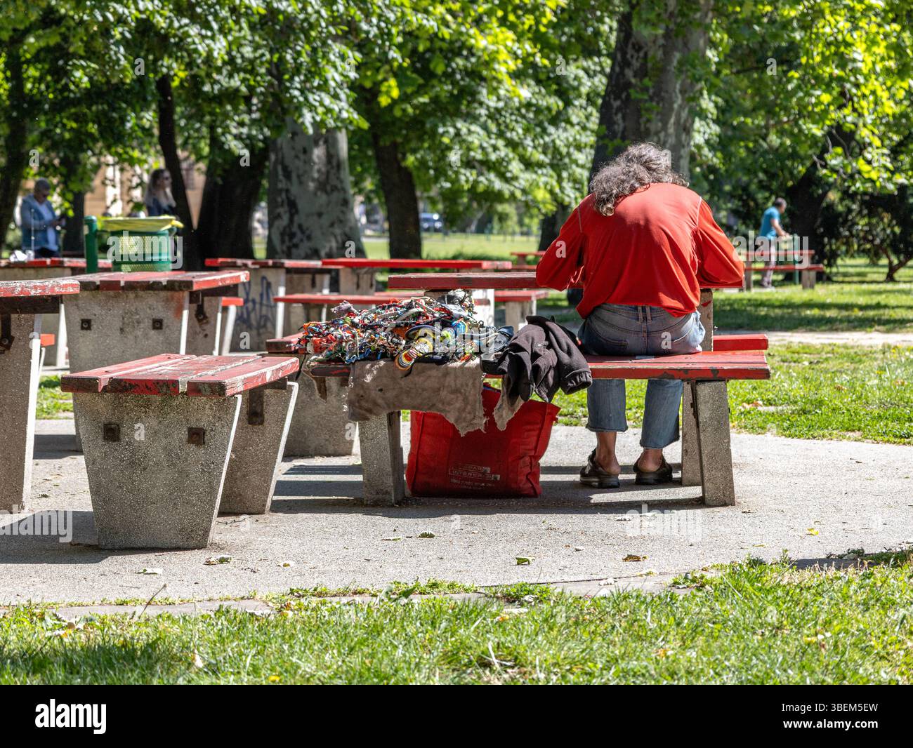 Uomo seduto da solo su una panchina in un parco pubblico con oggetti fatti a mano accanto a lui Foto Stock