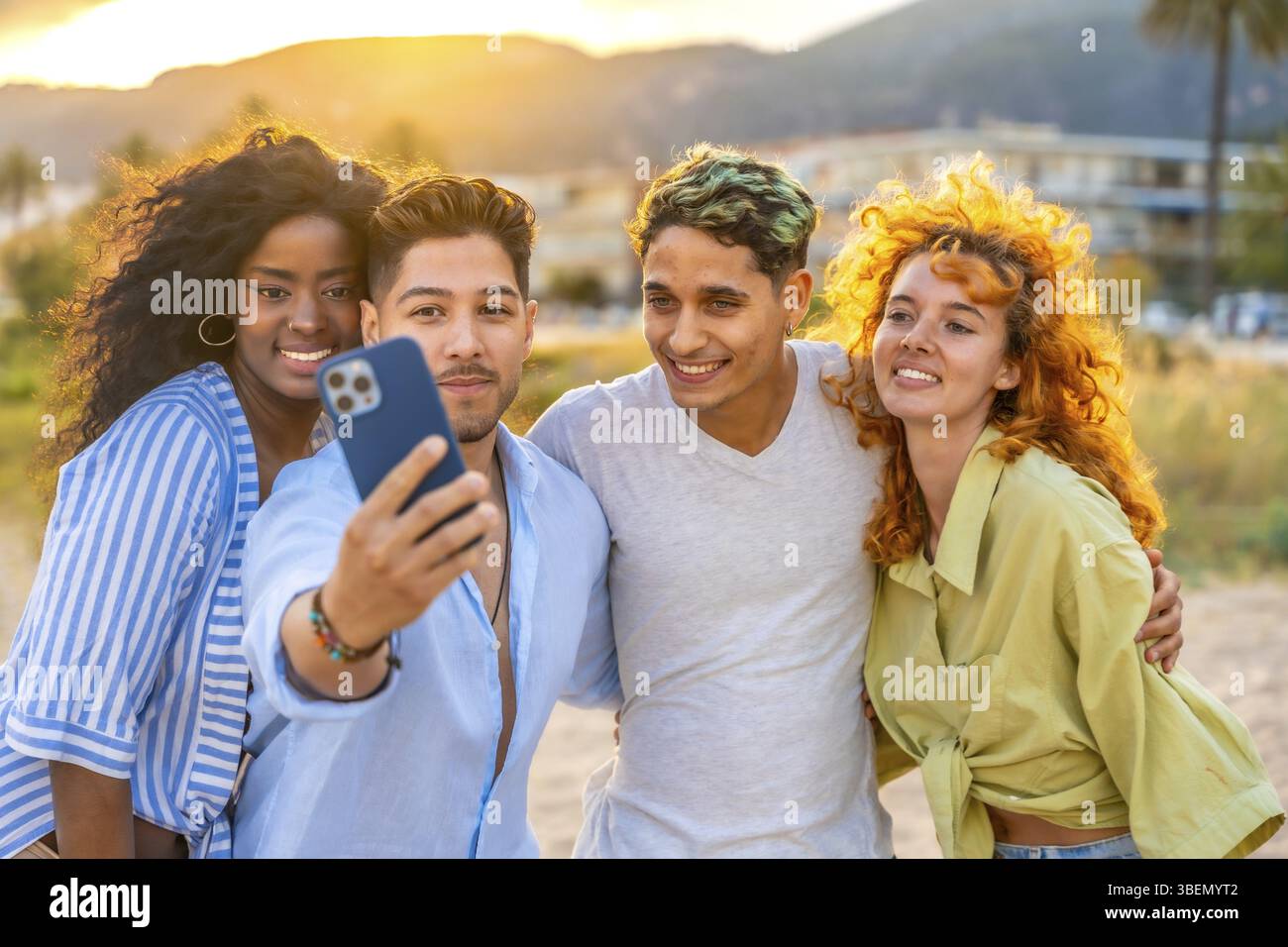 Quattro giovani amici allegri di sfondi diversi che catturano un gioioso selfie al tramonto su una spiaggia mozzafiato, creando ricordi duraturi Foto Stock