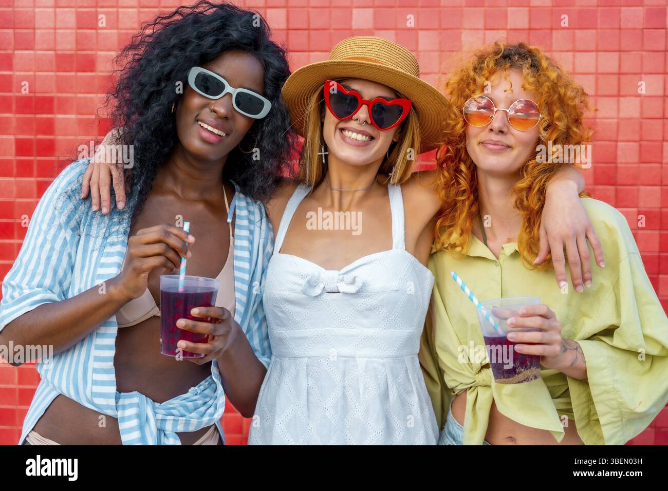 Tre giovani donne multietniche che si abbracciano a vicenda e bevono succhi di frutta con la paglia, appoggiate contro un muro di piastrelle rosse Foto Stock