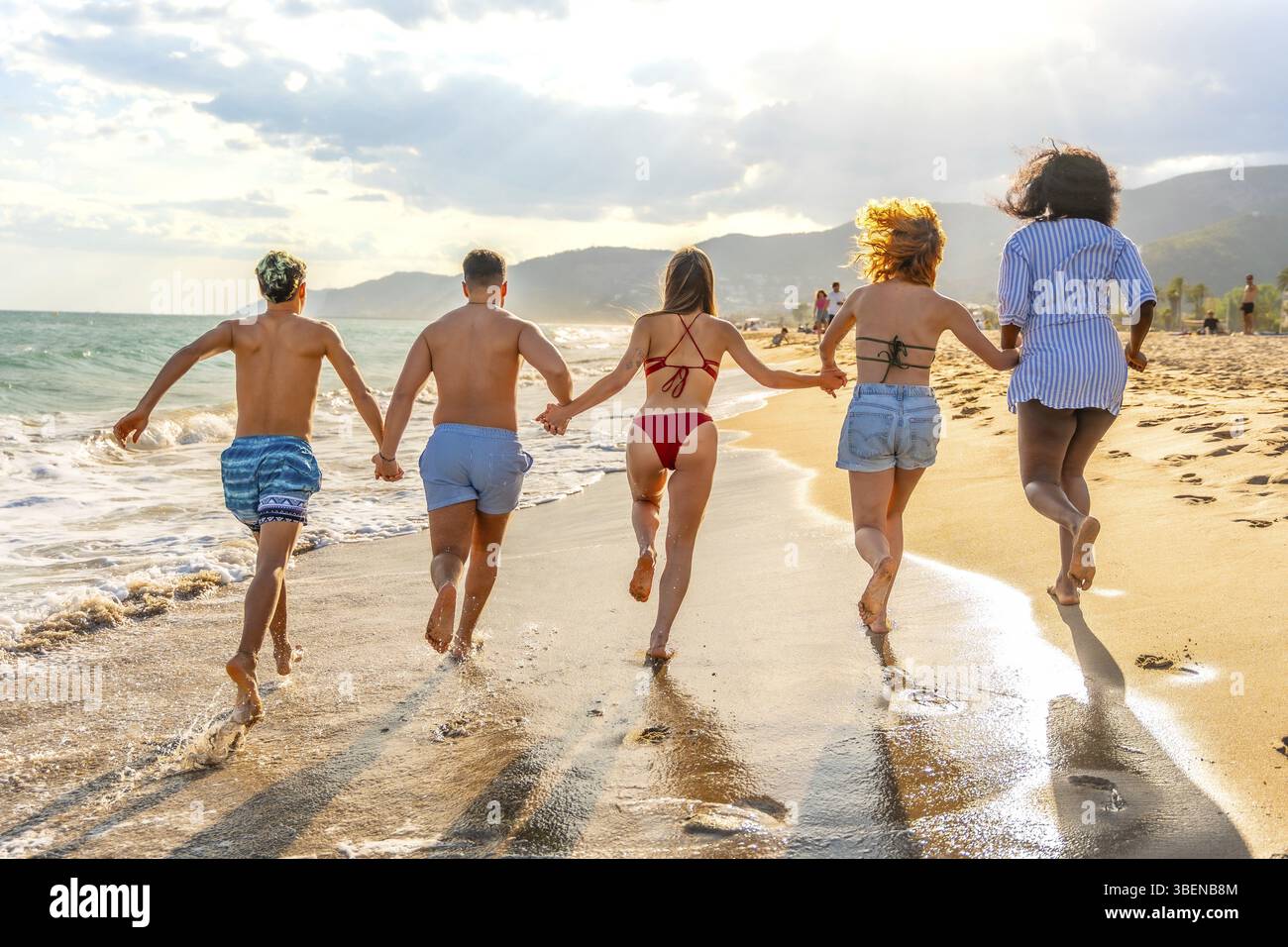 Cinque amici multietnici che corrono sulla spiaggia al tramonto tenendosi per mano e godendo insieme le vacanze estive Foto Stock