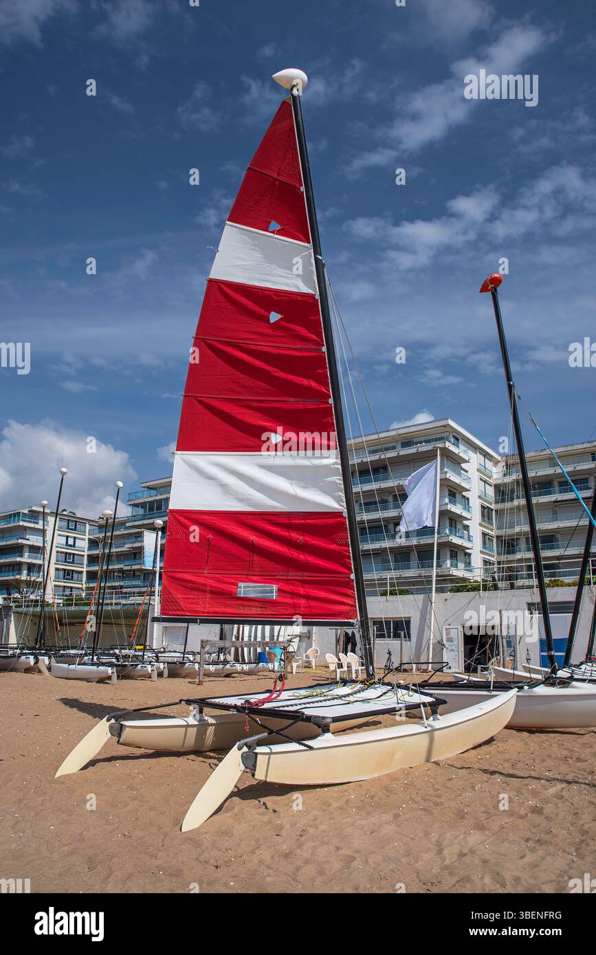 Un catamarano su una spiaggia sabbiosa in Bretagna, nella città di la Baule, con la sua vela rossa e bianca di fronte ad appartamenti Foto Stock