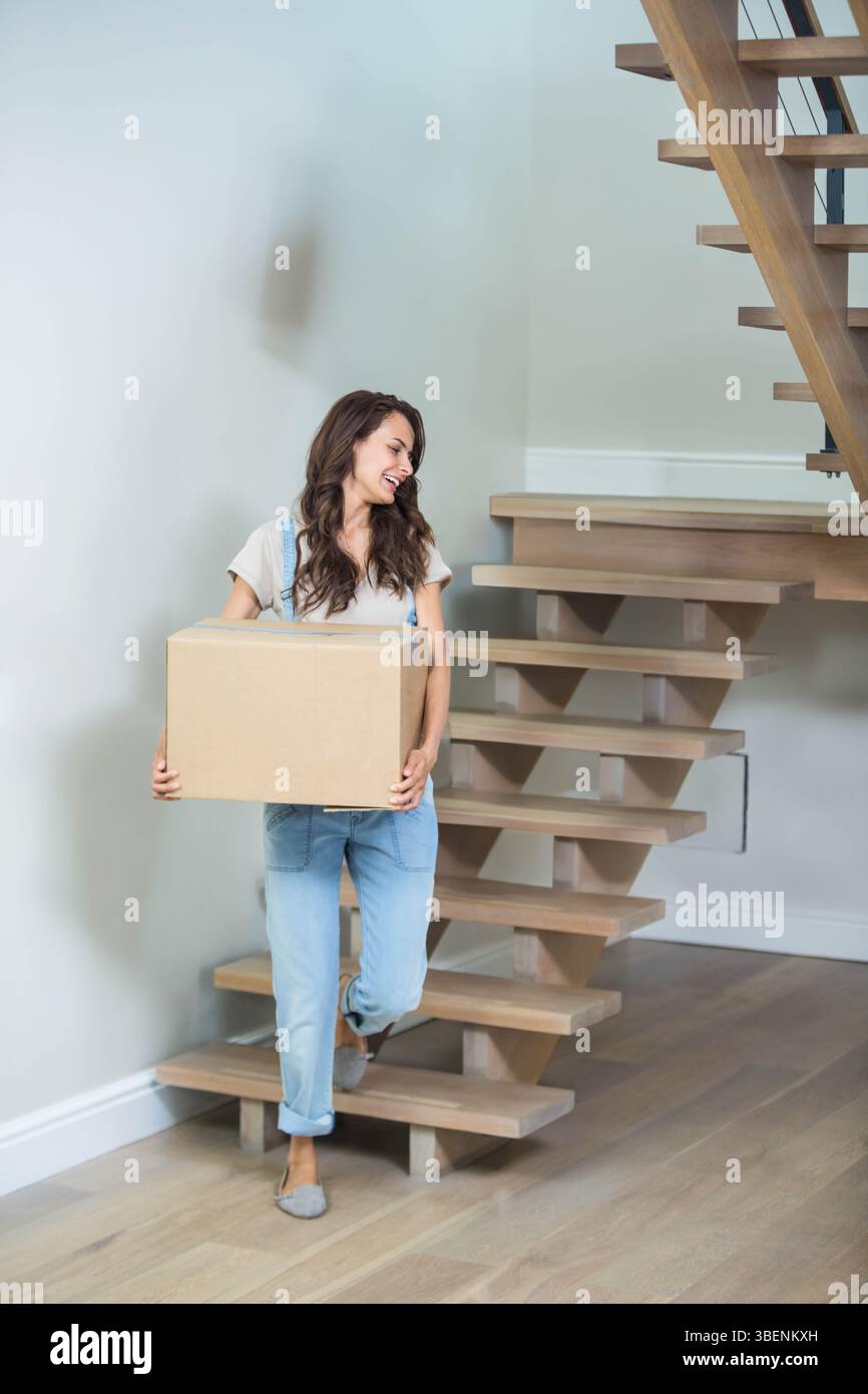 Donna adulta che porta una scatola mobile di cartone mentre sale la scala di legno nel vano scale di casa Foto Stock