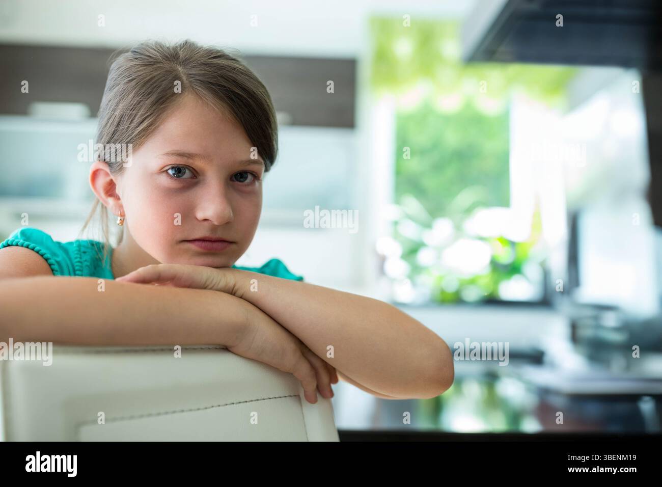Ragazza che indossa la parte superiore turchese appoggiata su una sedia bianca in cucina che mostra una finestra con fogliame sfocato Foto Stock