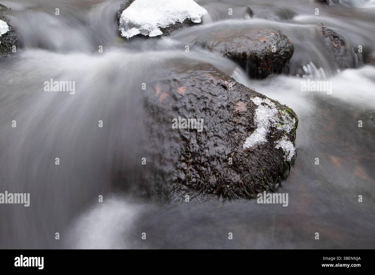 Rocce scure e lisce si trovano in un fiume invernale che scorre dolcemente, parzialmente coperto da sottili strati di neve e ghiaccio. Foto Stock