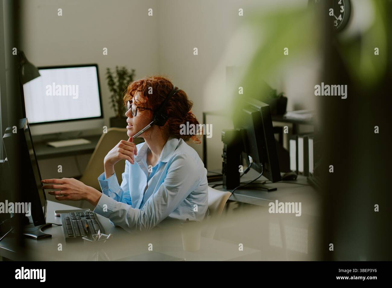 Donna seduta alla scrivania in un ufficio moderno, indossa la cuffia e parla sullo schermo del computer, con le attrezzature per l'ufficio in background Foto Stock