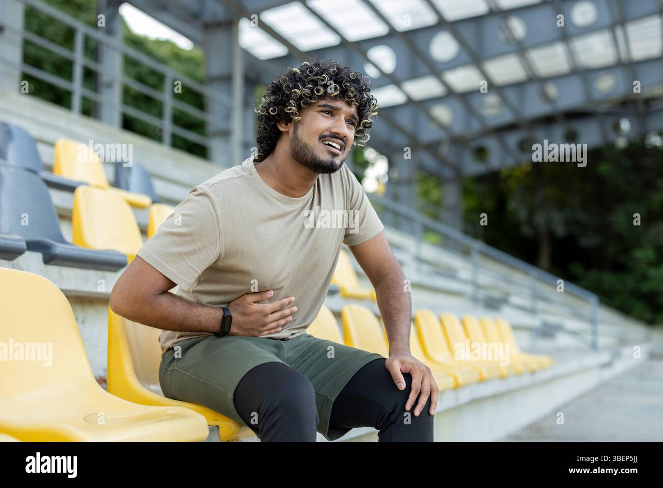 Un giovane atleta che soffre di disagio addominale mentre si siede sugli sbiancanti dello stadio dopo una sessione di allenamento all'aperto. Foto Stock