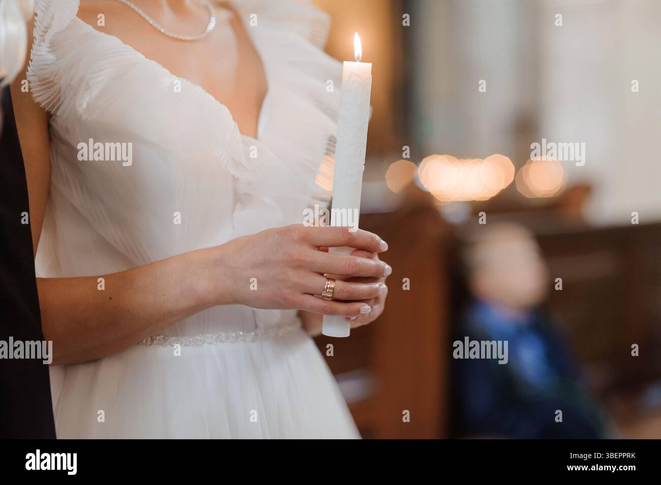 Una sposa radiosa in un abito bianco con volant tiene una candela accesa durante una cerimonia nuziale in un ambiente di chiesa, simboleggiando la fede e l'unità con un aspetto sfocato Foto Stock