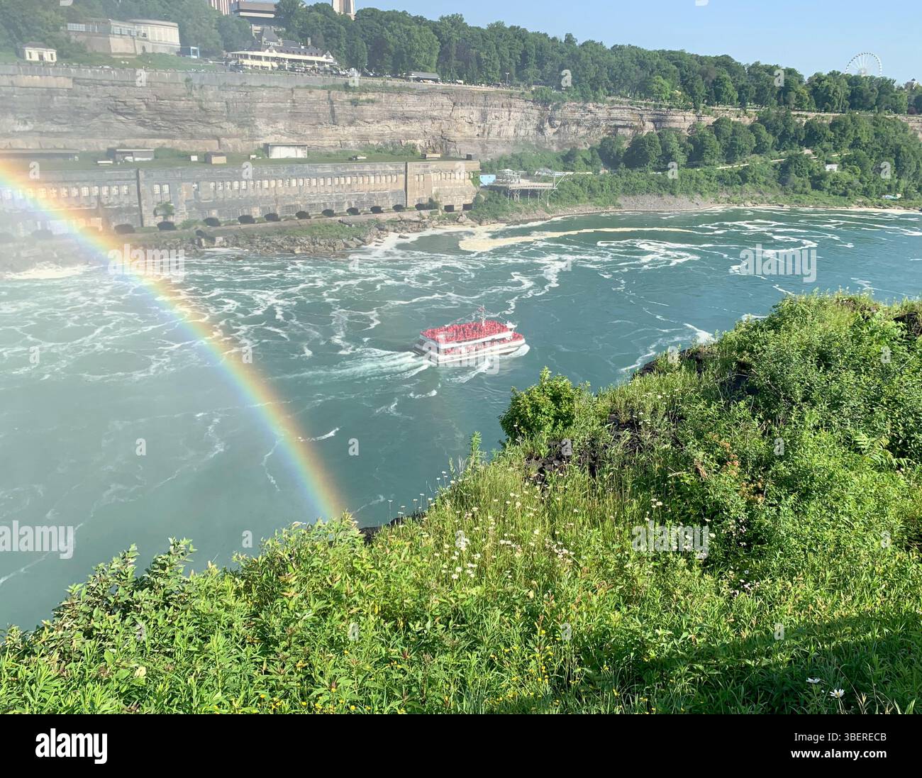 Il tour in barca attraversa il fiume Niagara sotto un arcobaleno vivido vicino alle cascate del Niagara in una giornata estiva di sole con le verdi scogliere dell'Ontario sullo sfondo. Foto Stock