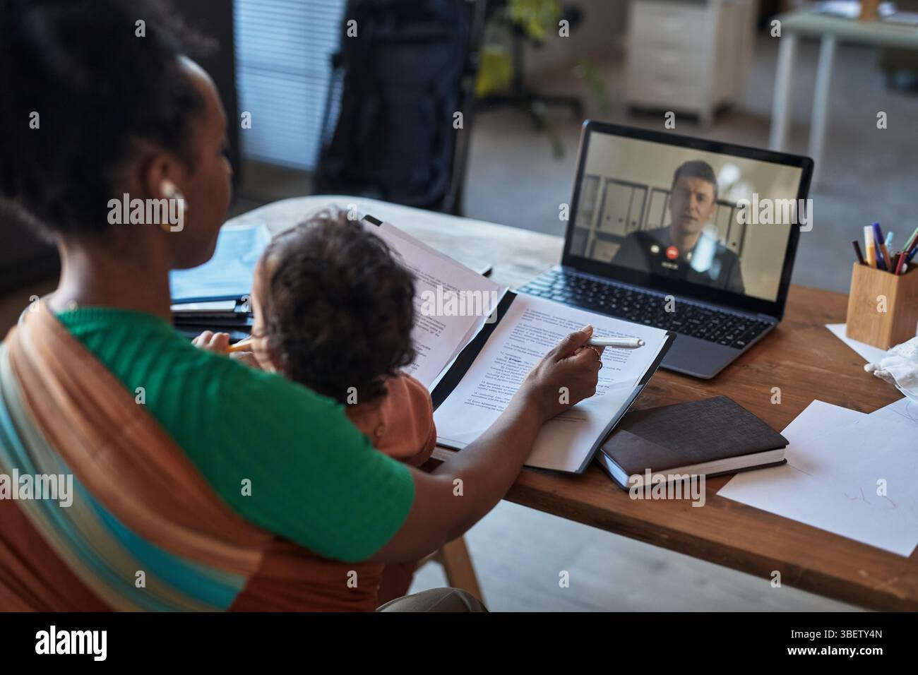 Donna che partecipa a una riunione video mentre equilibra le attività e tiene in braccio il bambino. Lo sfondo presenta l'impostazione dell'ufficio con documenti, laptop e blocchi note Foto Stock