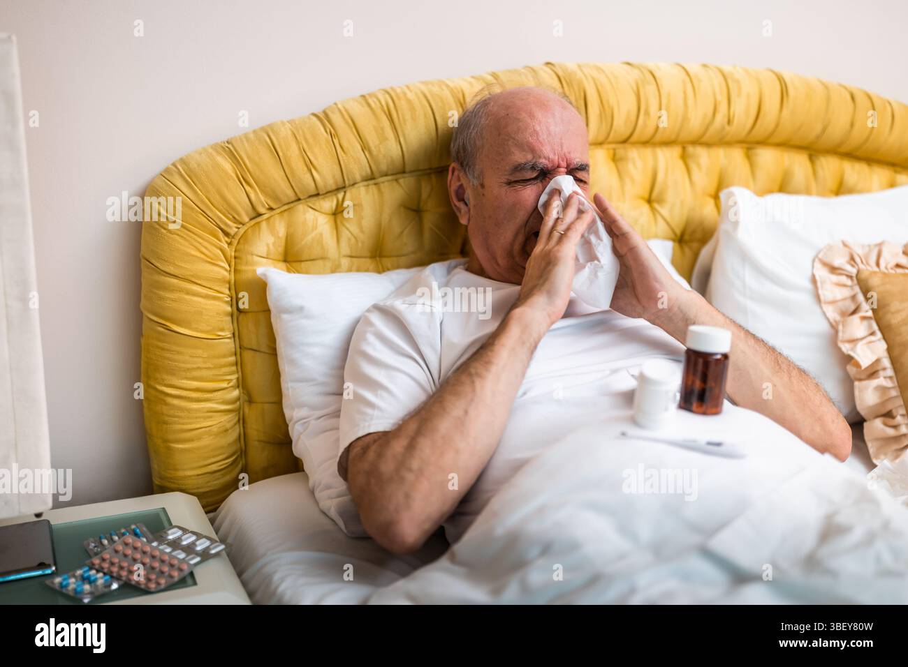 Anziano malato ed esausto che ha influenza e starnuti nel tessuto mentre sdraiato a letto circondato da farmaci a casa sua. Foto Stock