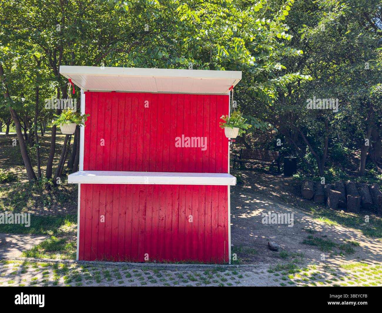 Piccolo chiosco rosso in legno con tetto bianco e vasi da fiori appesi, circondato da alberi verdi, luce del giorno estiva, ambiente esterno vuoto. Foto Stock