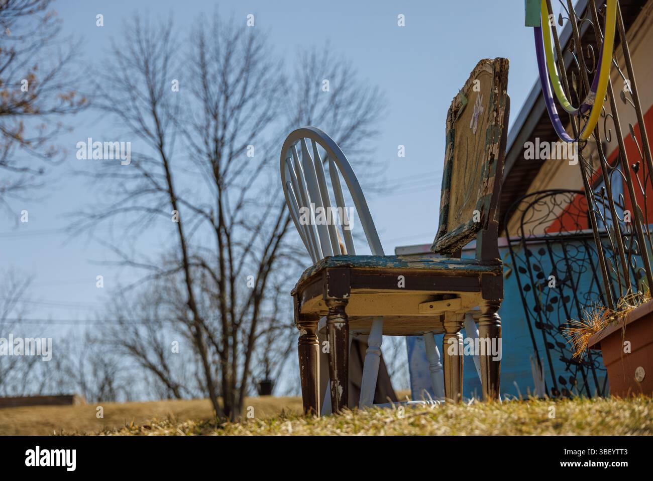 Due sedie non abbinate, una vecchia e intemprata, si siedono fuori da una casa colorata su un prato erboso all'inizio della primavera. Alberi senza foglie e un cielo blu limpido Foto Stock