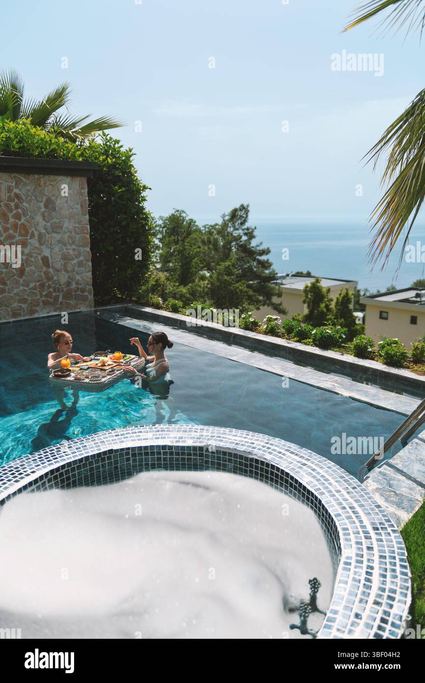 Una bionda e una bruna sui 20 anni che indossano costumi da bagno, gustate una colazione galleggiante mentre vi rilassate nella loro piscina privata. Foto Stock