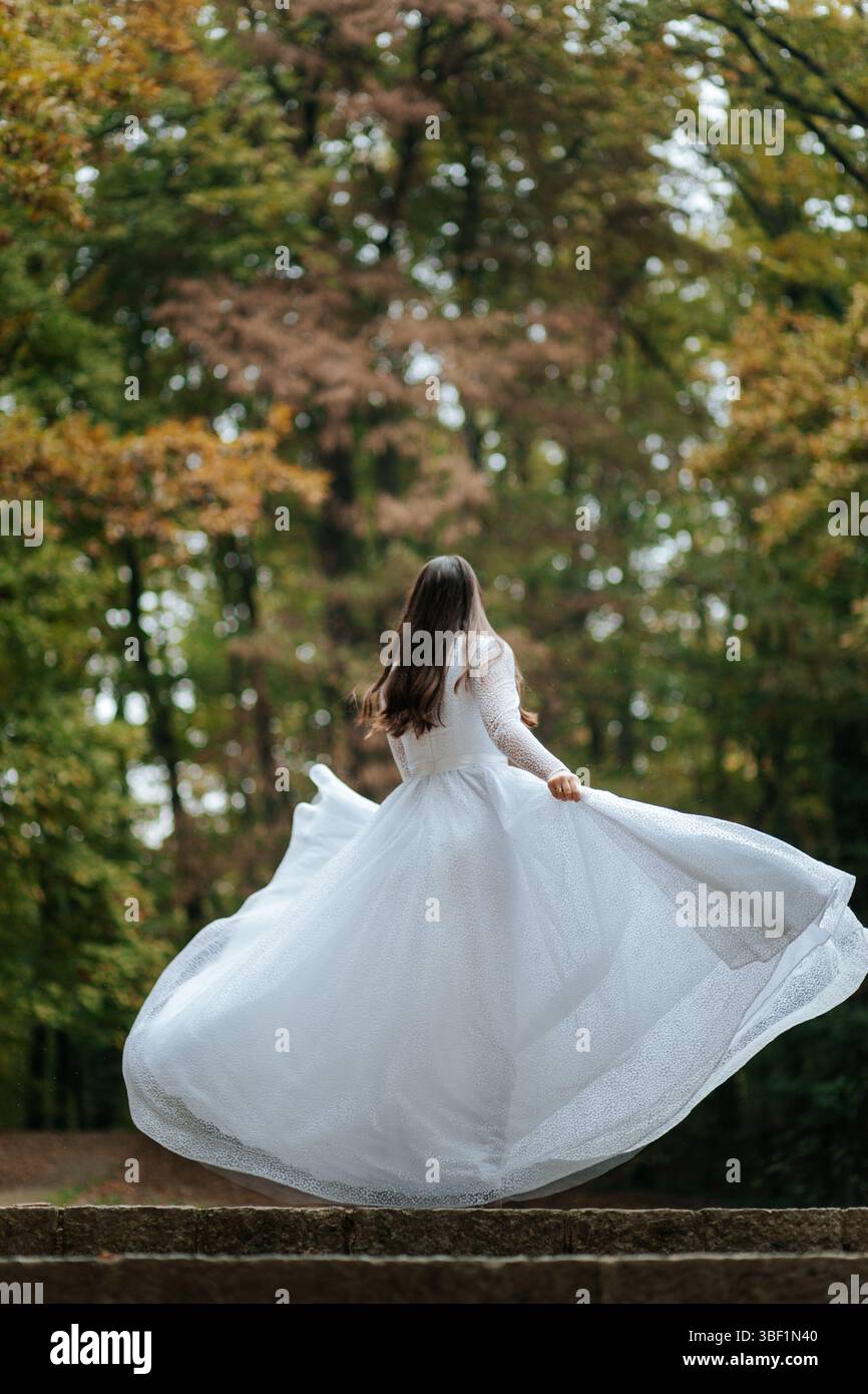 Una sposa si intreccia gioiosamente in una foresta, il suo abito fluente e il copricapo floreale aggiungono un'atmosfera eterea e incantevole. Foto Stock